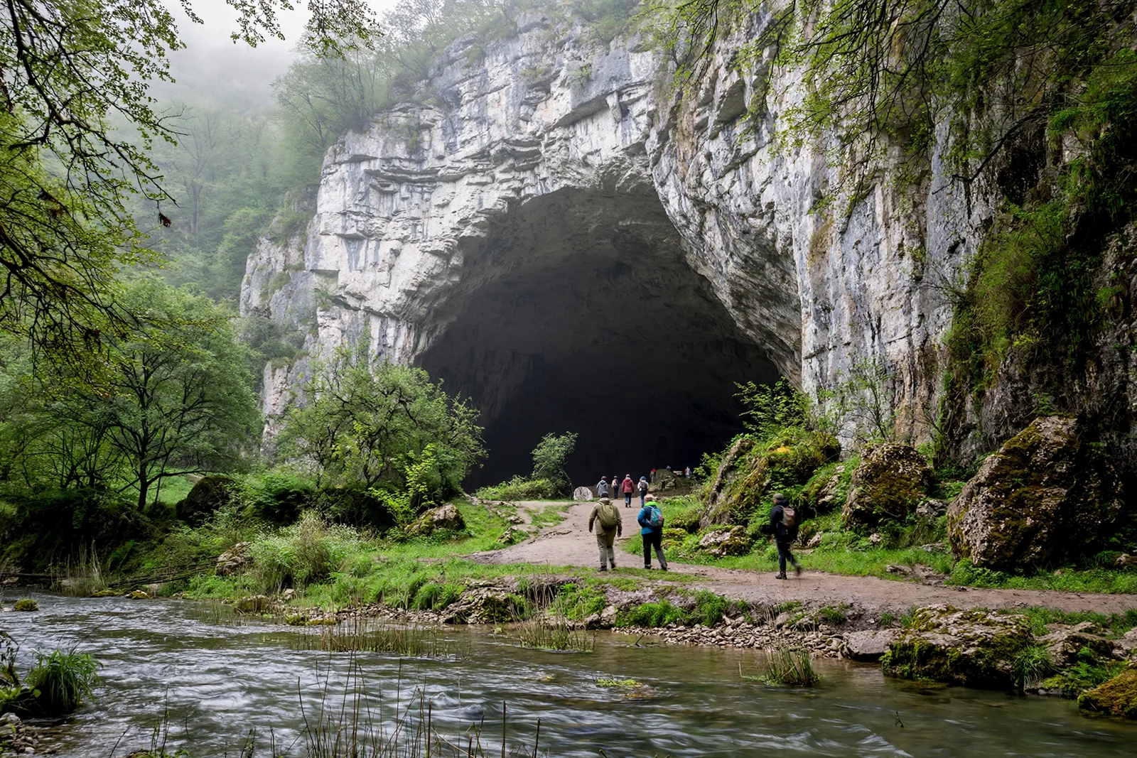 Huge arched entrance of Devetashka Cave in Bulgaria seen from the Osam river bank, with visitors walking toward the dark opening