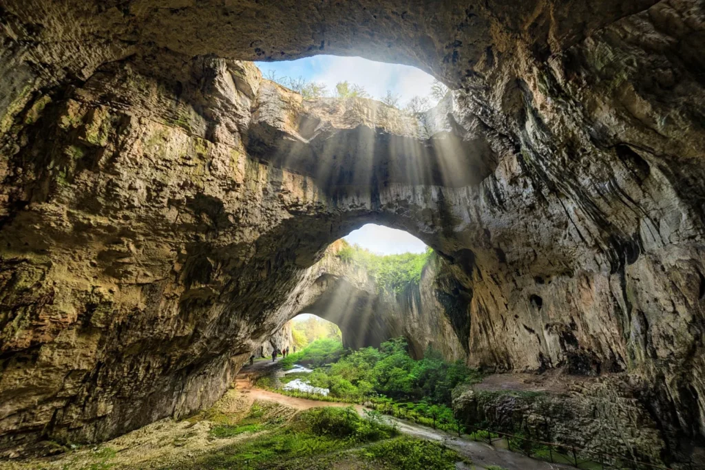 Sunlight streaming through seven ceiling openings into the vast hall of Devetashka Cave in Bulgaria, with a stream and green moss below